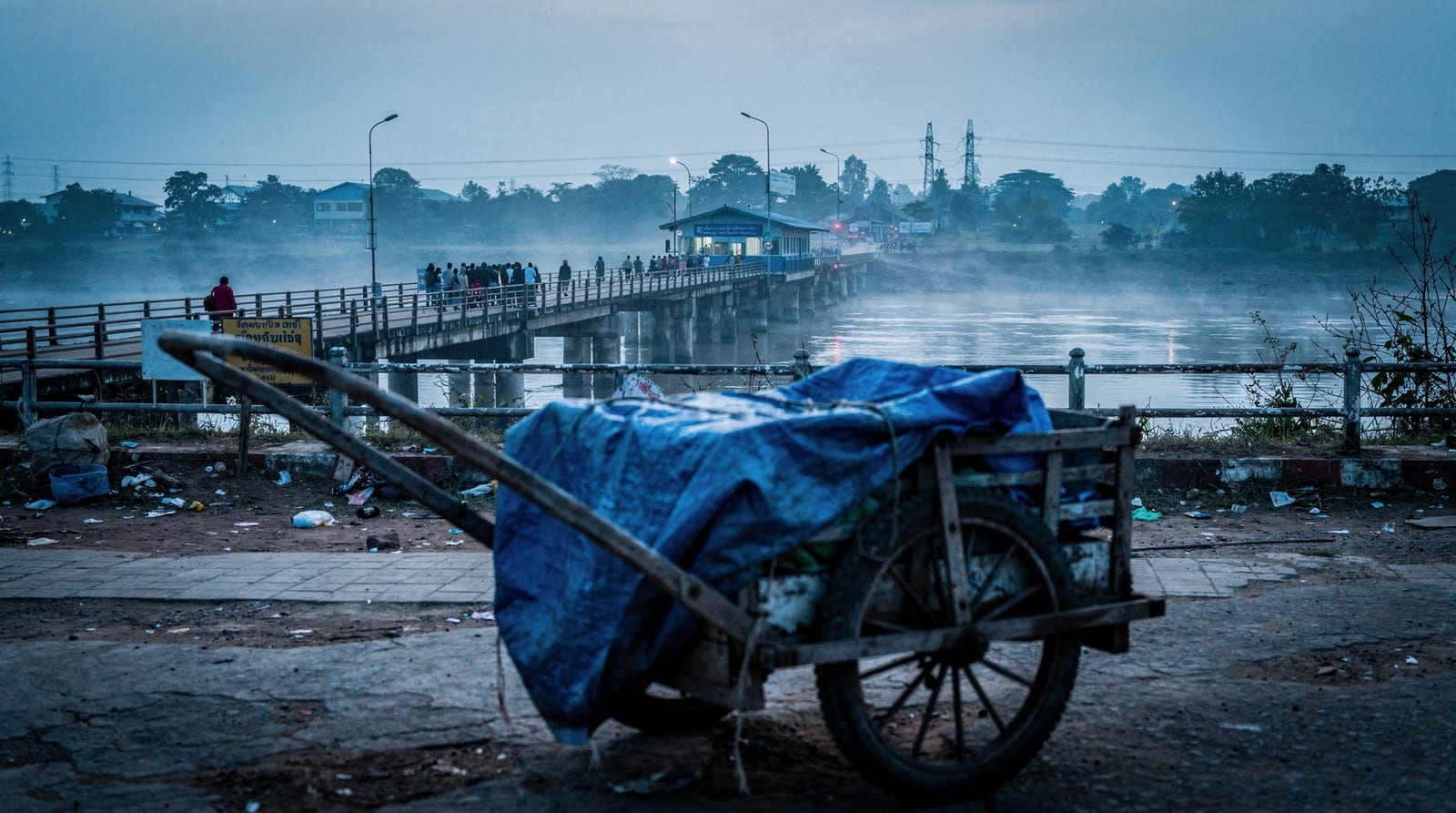 The Mae Sot border crossing at dawn, a hand-cart in the foreground and a queue forming on the bridge in river mist.