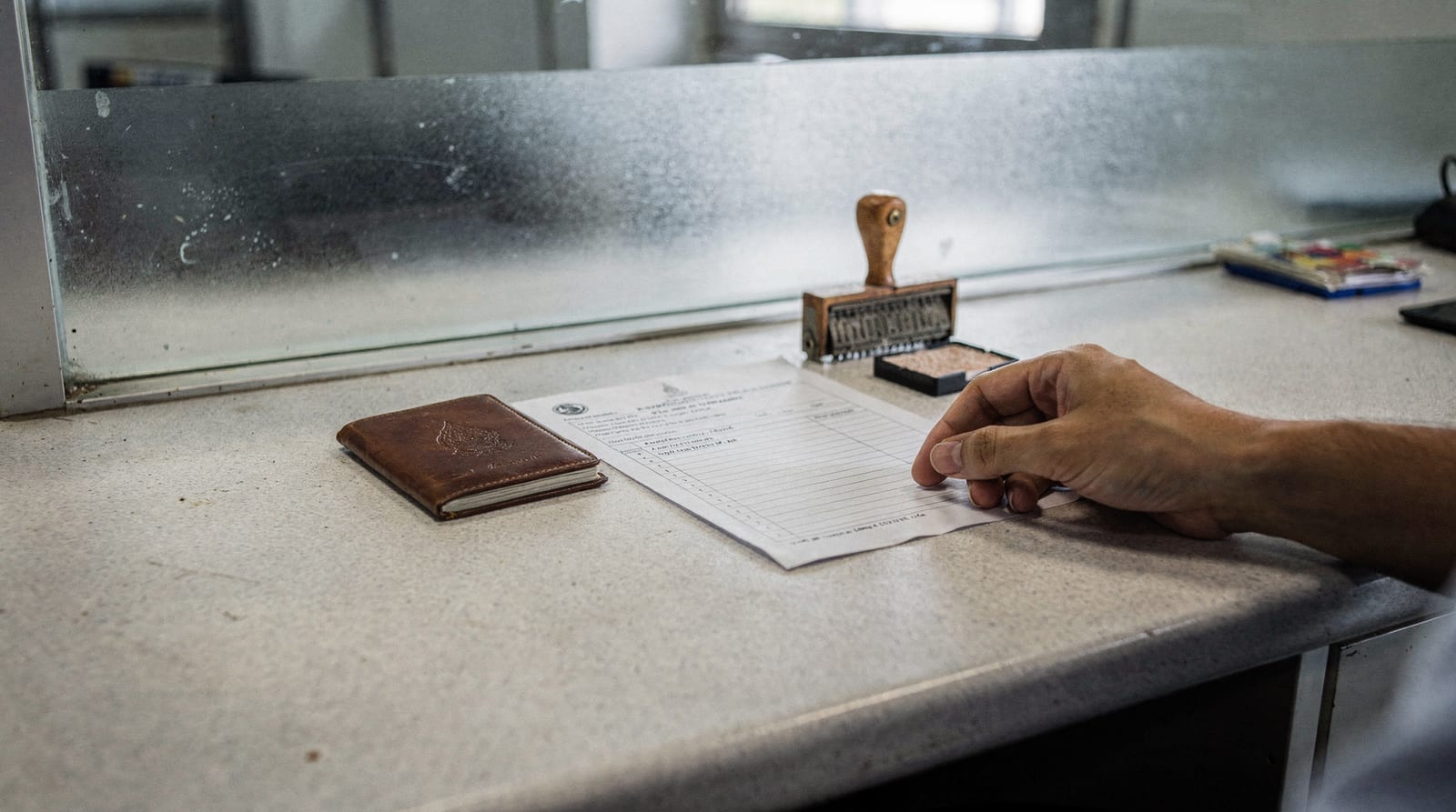 A generic passport, a blank application form, a date stamp and an ink pad on a Thai immigration counter, a hand sliding the form forward, no face.