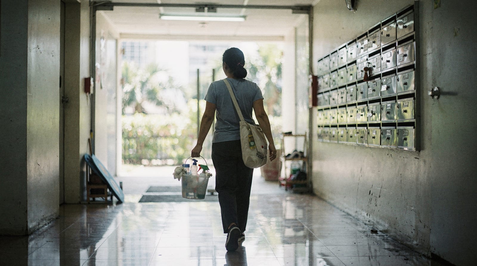 A day maid, seen from behind, walks through a Bangkok condominium corridor carrying a bucket of cleaning supplies and a tote bag.