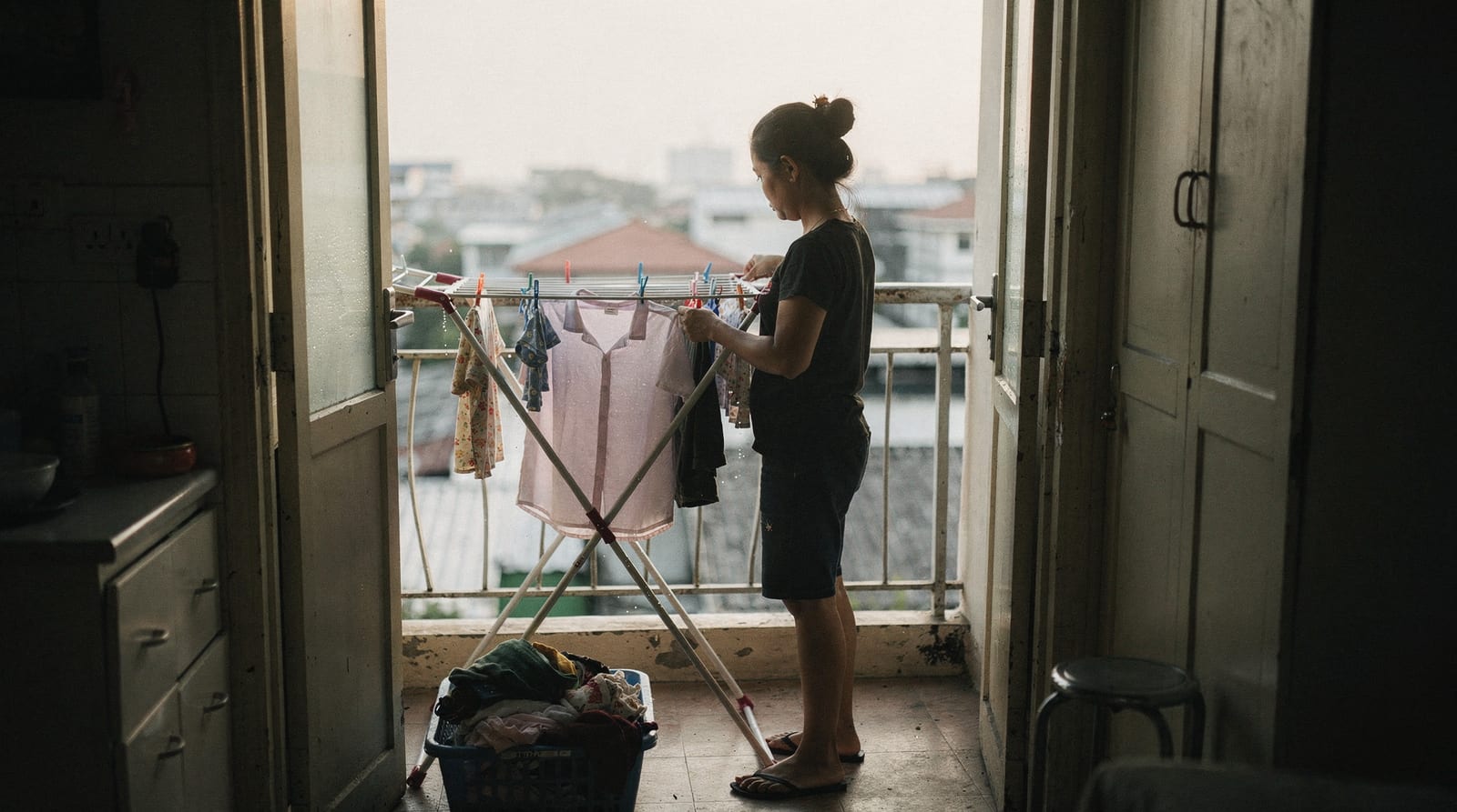 A live-in domestic worker, seen from behind in early morning light, hangs washed laundry on a folding rack on a small Bangkok service balcony.