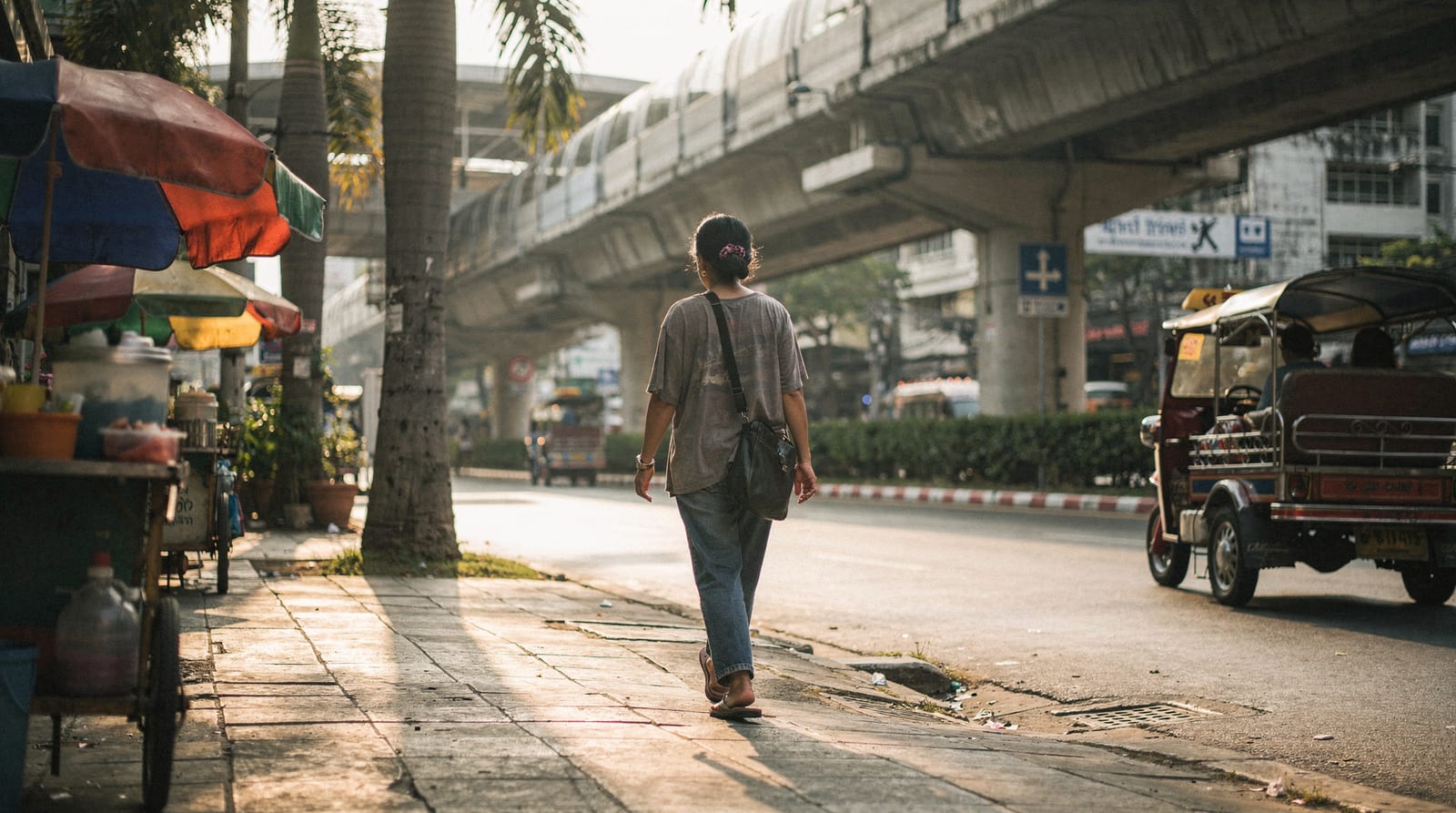 A domestic worker in off-duty clothes, seen from behind, walks toward a Bangkok BTS station on her day off, a street vendor cart and tuk-tuk nearby.