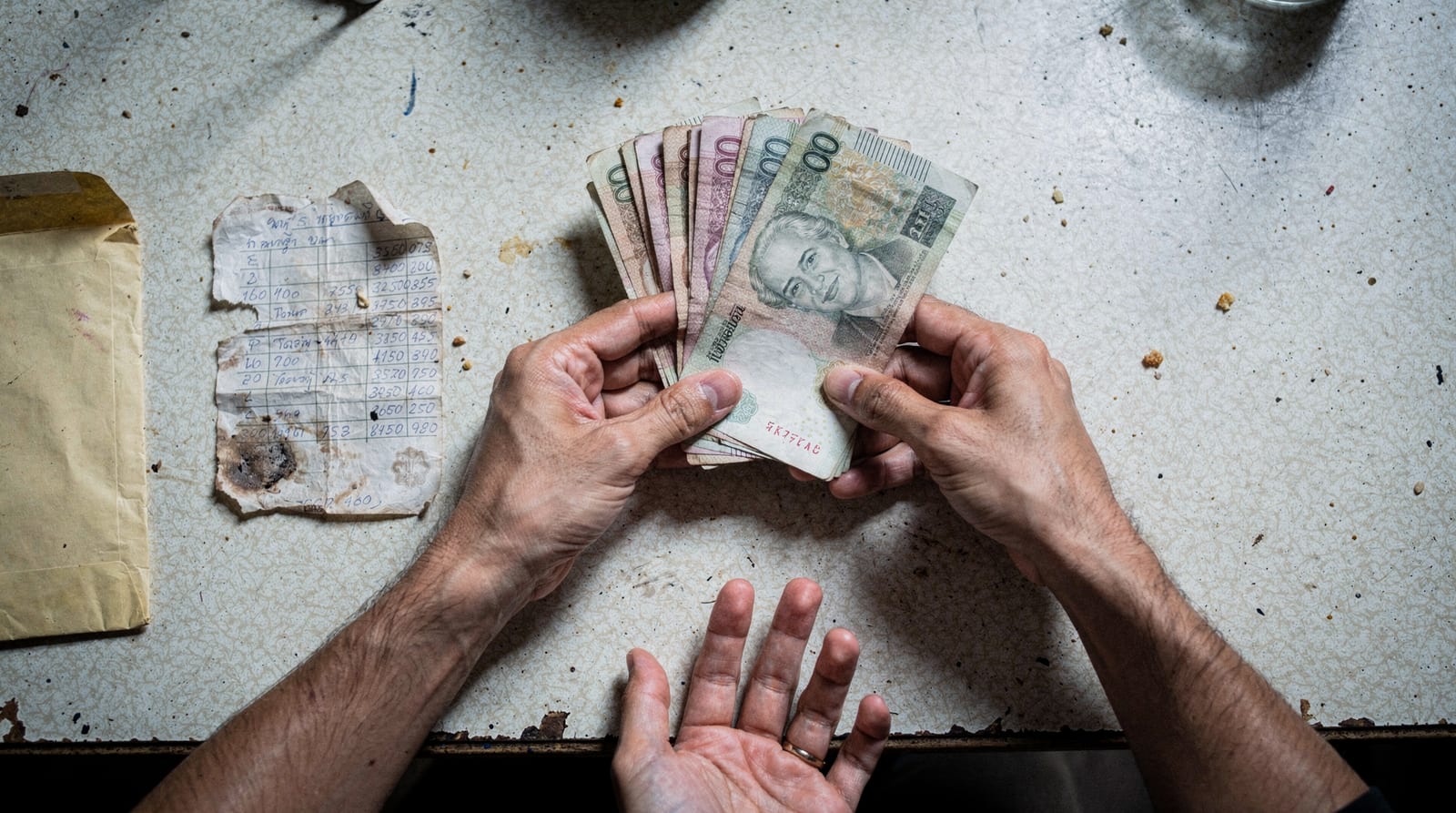 Hands counting baht banknotes beside a torn document and envelope, showing money taken as a placement fee.